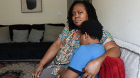 Black woman (mom) in print blouse holds a young Black boy in a light blue T-shirt, both sitting on a gray couch in a living room 
