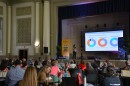 People sit around tables, watching a woman on a stage give a presentation.