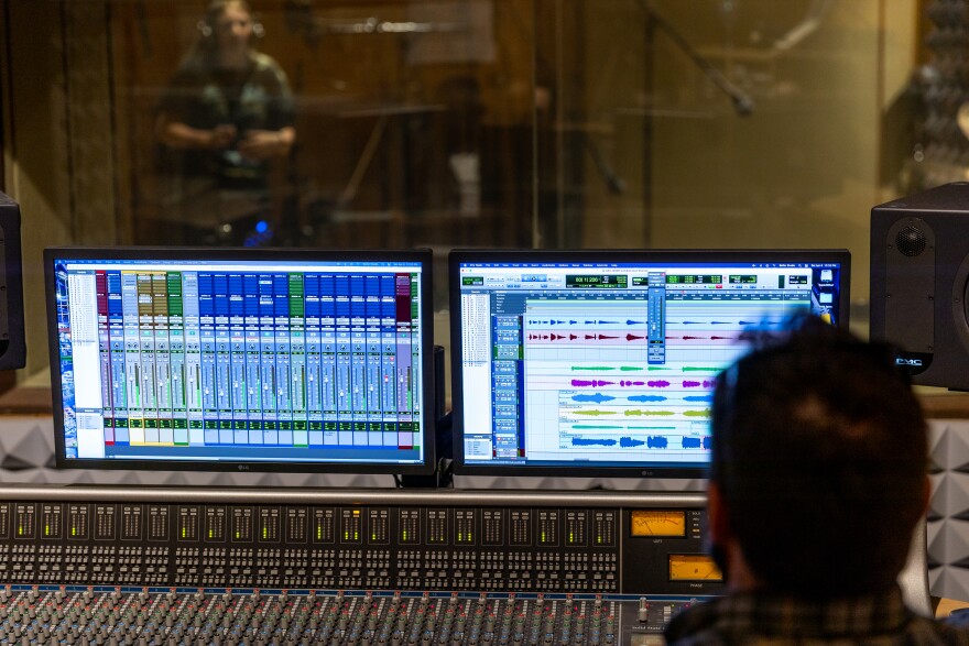 A musician, seen behind a glass window, takes direction from a producer seen in the foreground, sitting in front of an audio mixer in a recording studio.