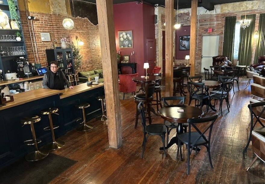 Tables and chairs inside Pausa Coffee in Syracuse, with wood floor and brick walls behind a bar  