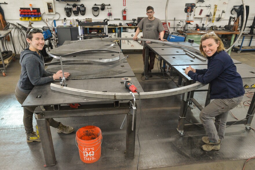 Three people, two women and a man, stand around several large, steel tables that hold different metal parts for a large sculpture.