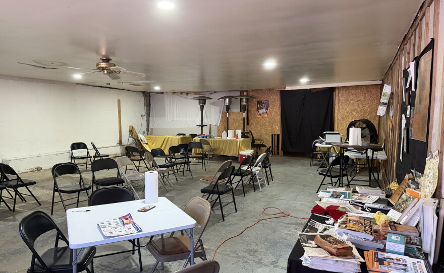 A large room with concrete flooring. About two dozen metal chairs are in the center of the room. A small white table is in the left bottom corner. There are long tables against the wall. The back and left wall is covered with wooden panels.