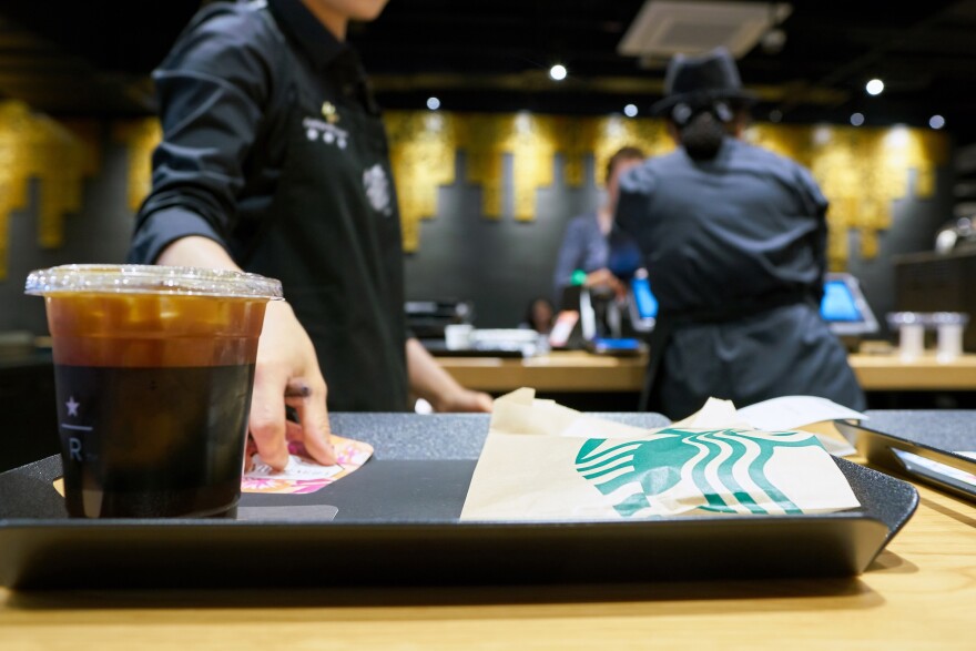 In the foreground, an iced coffee and paper pastry bag with Starbucks branding sit on a tray. A barista is placing a bit of paper on the tray. Behind them, another person works a cash register.