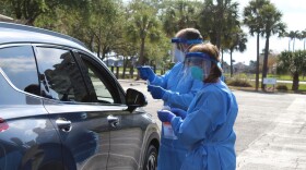 2 people in medical scrubs and personal protective gear collect specimens from a car at a COVID-19 drive-thru testing site.
