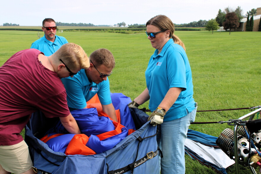 Four people pack a colorful hot air balloon into a large blue bag on a grassy field. Two wear matching blue shirts and gloves, working together while another stands nearby watching. Equipment is visible in the corner.