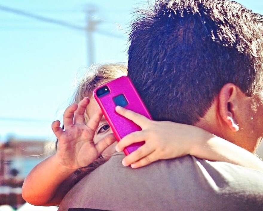 A little girl looks over her father's shoulder at a smartphone.