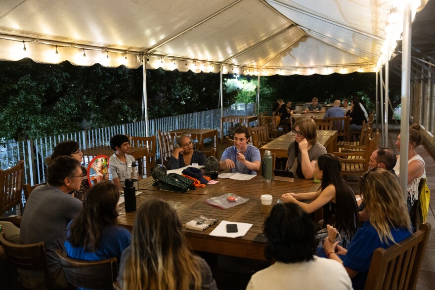 A group of people sit around a table and listen as one person talks. 