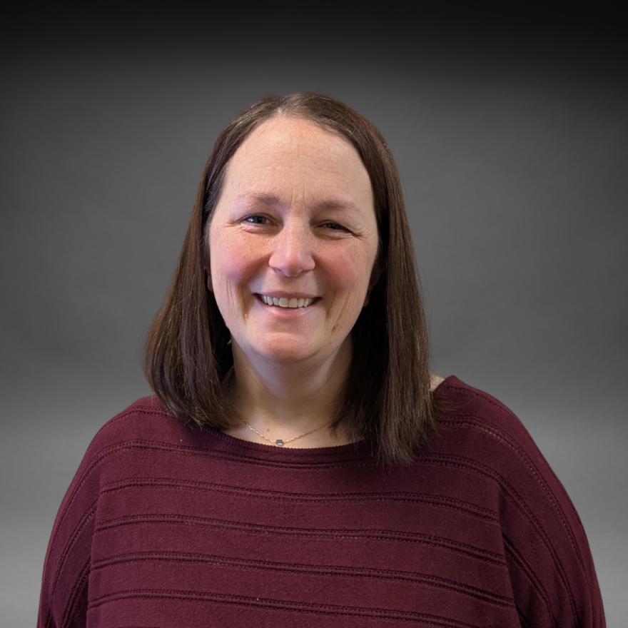 A woman with straight brown hair, wearing a maroon sweater, smiles at the camera in front of a dark gray background.