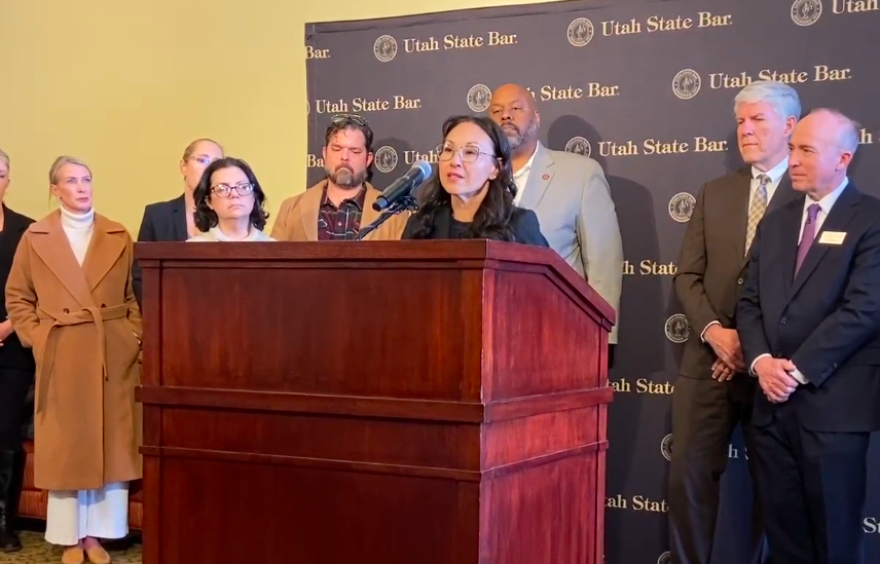Kim Cordova, president of the Utah State Bar, speaks from behind a podium at a press conference. Members of the Bar stand behind her in front of a backdrop reading “Utah State Bar.”