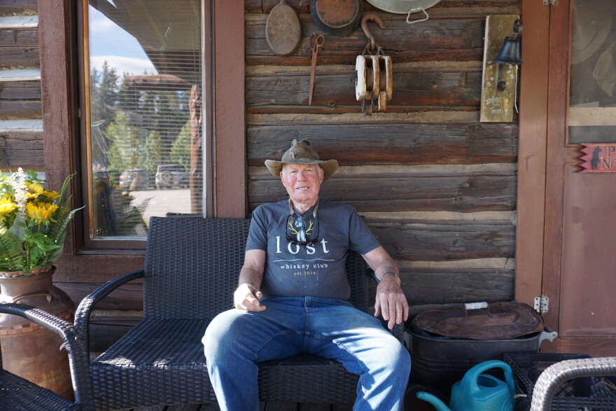 An older man sits on a wicker bench outside a wooden cabin. He’s wearing a western hat and his shirt says “lost.”