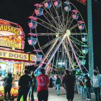 Ferriss wheel in neon lights with people in the foreground walking around wearing masks.