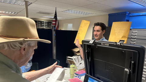 An older man wearing a tan fedora holds a long slip of paper. A younger man standing across a desk from him holds up two large envelopes of election related documents.
