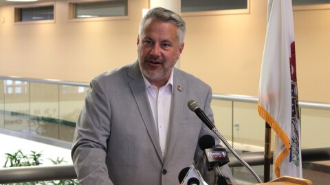 A man in a grey suit stands in front of microphones while giving a speech.