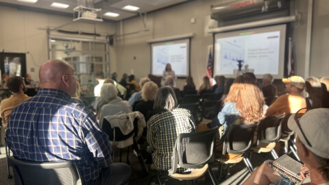 People sitting in a large room listening to a woman speak with two projector screens behind her