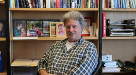 A man sits in front of a bookshelf full of textbooks and photos and smiles. 