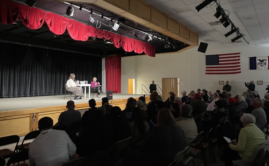 Two women sit on a stage in front of a large crowd in an auditorium