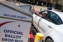 A person leans out of their car in a parking lot to drop a ballot into a drop box, labeled Salt Lake County Official Ballot Drop Box.