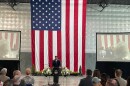 Mayor Wagoner stands a podium in front of a very large American flag hanging behind him, flanked by two large screens, and speaks in front of a seated crowd. 