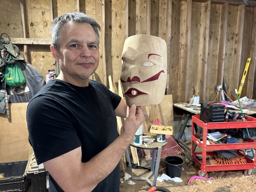 Micah McCarty stands in his workshop wearing a black T-shirt and holding up a nearly-finished mask that is carved from cedar and painted with red lips and eyebrows. It has abalone-inlaid white eyes. He is grinning an proudly displaying his ar. The workshop is full of sawdust and jumbled tools are everywhere.