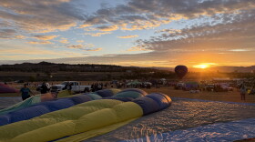 A hot air balloon lays deflated on the ground with the sunrise in the distance. The balloon lays on an open field with a sparse crowd in the background.