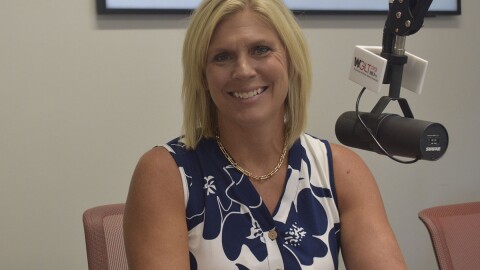 Woman in radio studio wearing flower-patterned blue and white sleeveless top and seated next to a microphone 