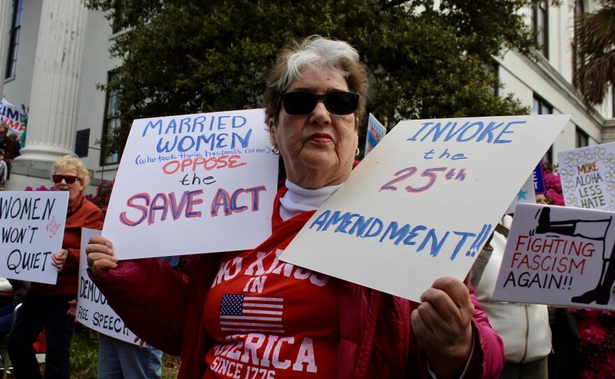Protestor holding two signs outside of Thalian Hall during the No Kings protest. The left sign states "Married women (who took their husbands last name) oppose the Save Act" and the right sign states "Invoke the 25th Amendment!!"