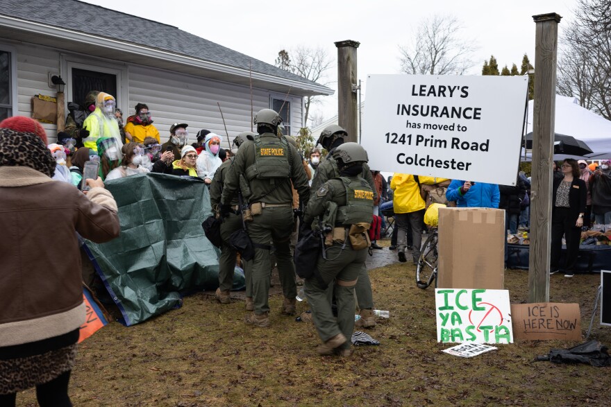 Officers from Vermont State Police approach protesters in face masks outside a home.