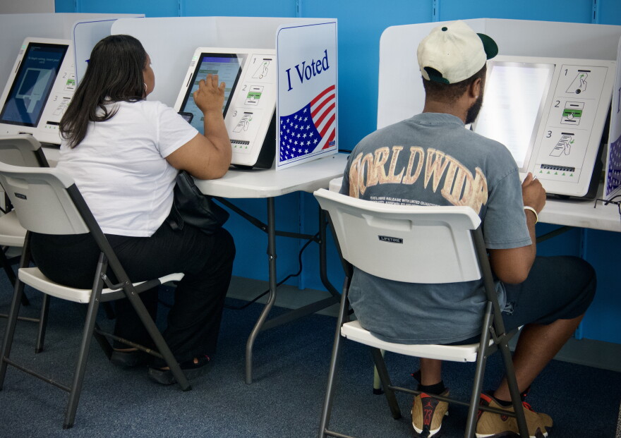 Interior photo that shows two people sitting in folding chairs with their backs to the camera. They appear to be making selections on electronic screens that are voting machines.