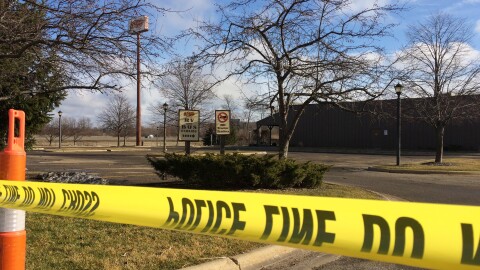 Police tape surrounds a black building with an empty parking lot. The grass is short and the trees are bare, denoting the winter season despite the lack of snow. The sign for Cracker Barrel can be seen at the top right of the image, partially covered by the branches of a tree in the foreground. 