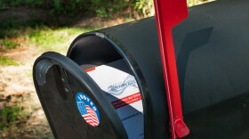A black mailbox with an "I voted" sticker on it, slightly ajar. There is a mail-in ballot inside and the red mailbox flag is up.