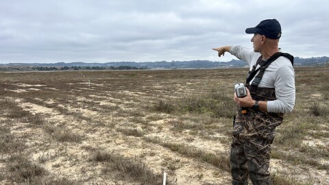 A man wearing boots and waders stands in a marsh field holding a gun-shaped machine in one hand while pointing towards the background, where there's a slough.
