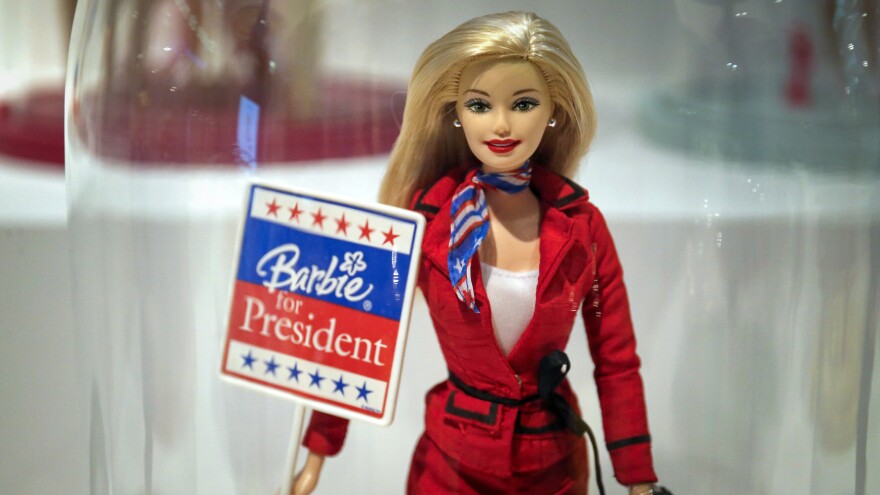 Photograph of a Barbie doll wearing a red blazer and red, white, and blue scarf holding a "Barbie for President" sign. 