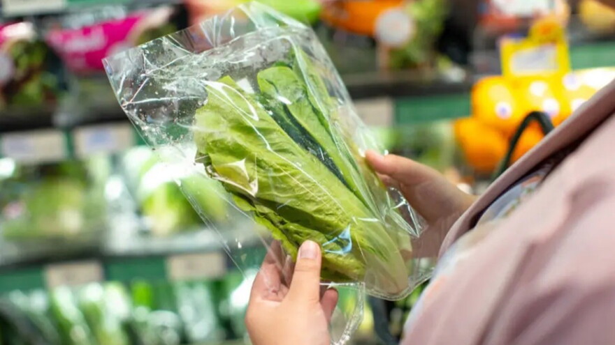 Fresh lettuce in plastic packaging held by shopper in grocery store, surrounded by various vegetables.  