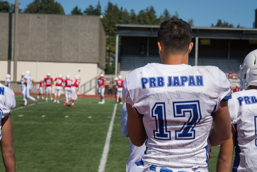 The back of a Japanese high schooler wearing white football gear standing on the sidelines of a football game. The jersey reads, "PRB Japan, 17."