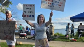 A woman wearing a white shirt and muted yellow floral skirt is holding a sign that says "Trump for Prison 2025" in her left hand and other sign in her right. There are other people and signs to her left and back.
