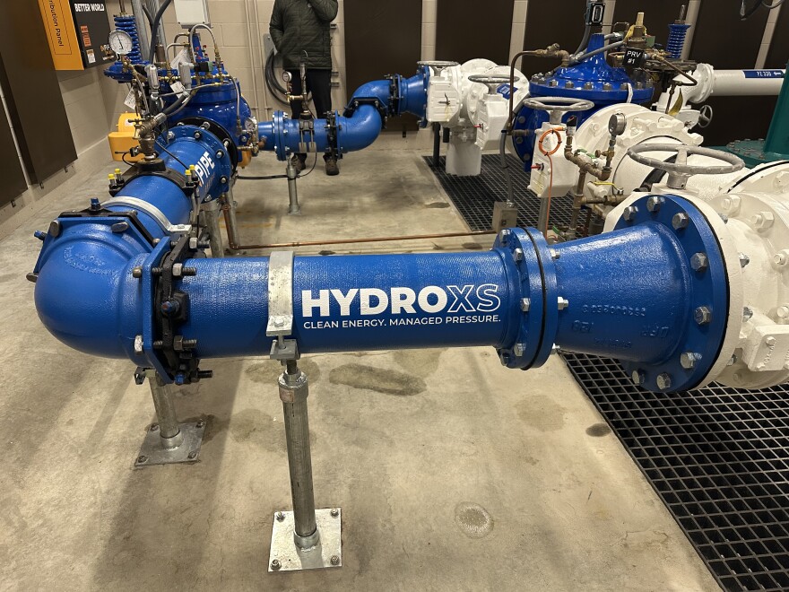 The curved blue pipe that holds a micro-hydropower turbine is shown inside a pumphouse at Skagit PUD, with the logo 'HydroXS' painted in it in white.