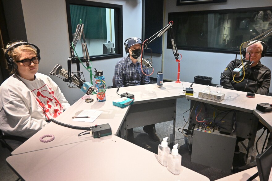 Three people sit inside a radio studio at tables with microphones in front of each one.