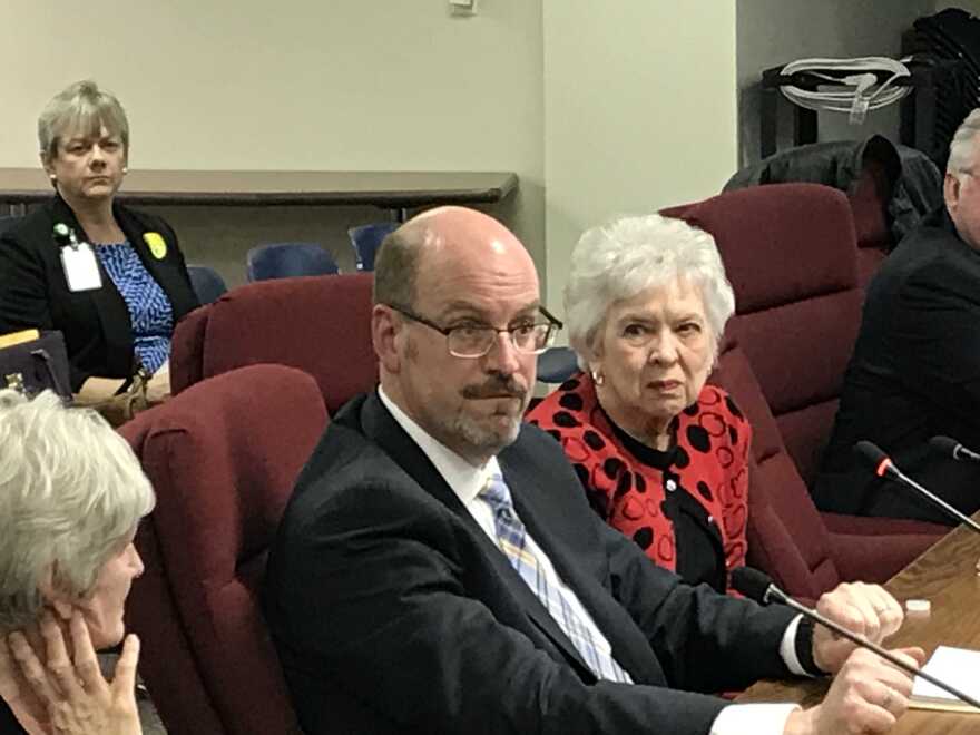 A man and a woman sit in chairs talking to elected government officials for McLean County, Illinois.