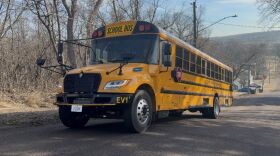 A yellow school bus is climbing a hill. There are many trees without leaves to the left of the bus.