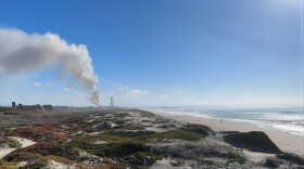 A blue sky day with sand dunes next to a beach. Smoke is drifting up from a fire in the background. The fire is faintly visible in the distance next to the two smoke stacks at the Moss Landing Power Plant