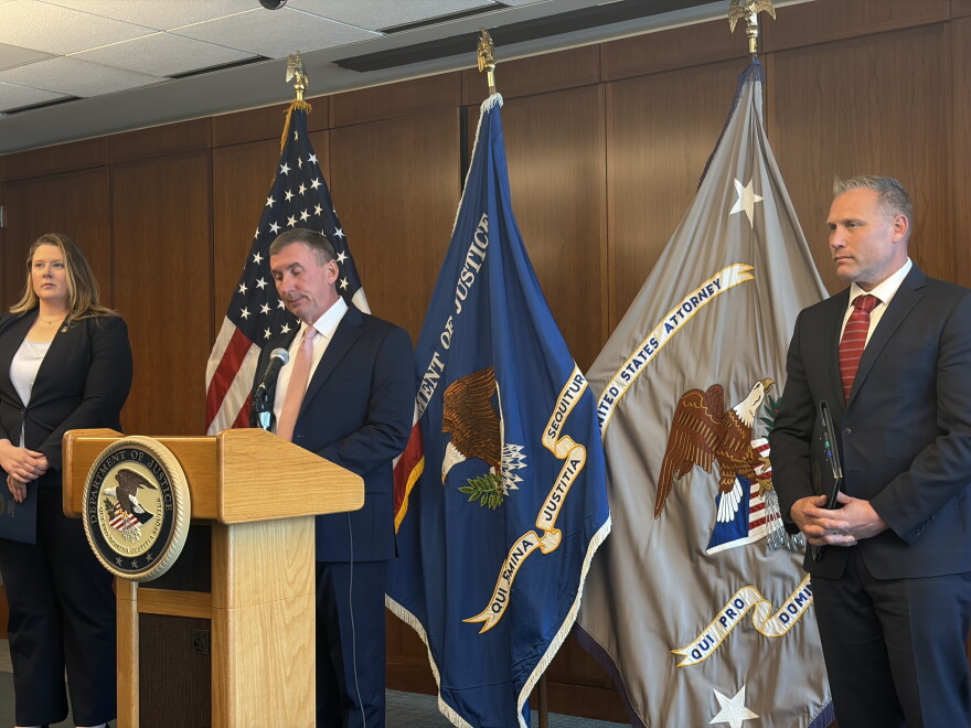 Federal officials stand in front of flags at a podium at a news conference. The podium has the Justice department's logo on it. 