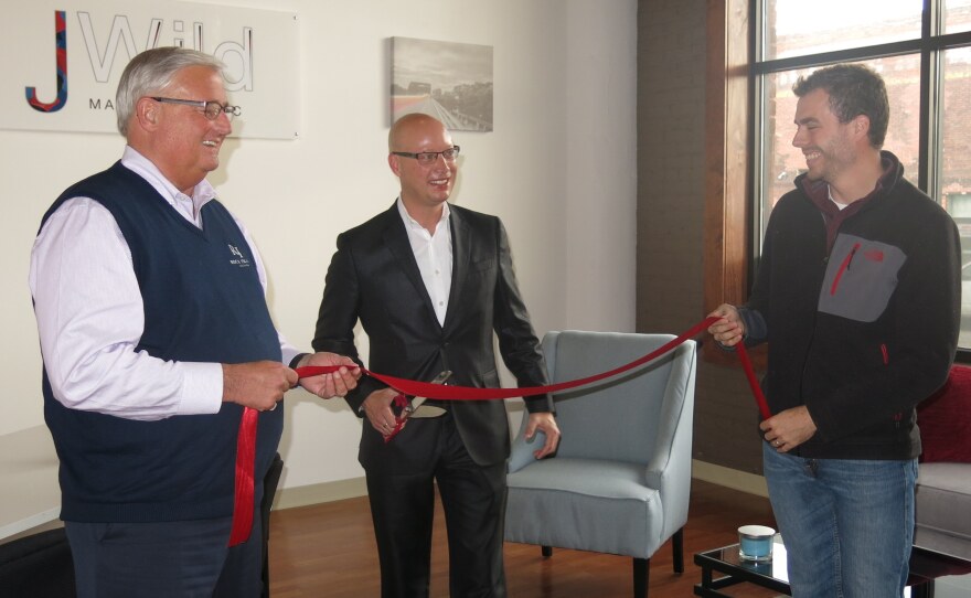 The mayor holds one end of a red ribbon while owner, Jason Wild, holds a large pair of scissors to cut it.