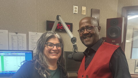 On the left, Mona Seghatoleslami smiling wearing a green dress and black jacket. On the right, viola professor George Taylor, also smiling, wearing a black shirt with a red vest. They are standing in a studio in front of a microphone and a computer.
