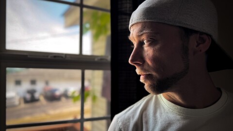 A man wearing a white tee shirt and backwards hat looks through a window.