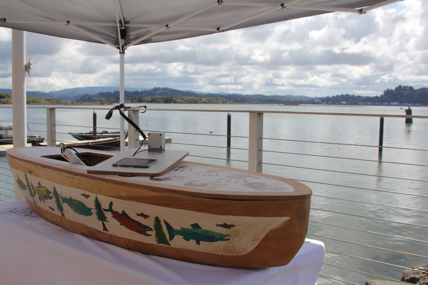 A small wooden canoe decorated with paintings of trees and fish on a display table set up on a boat ramp overlooking a bay. 