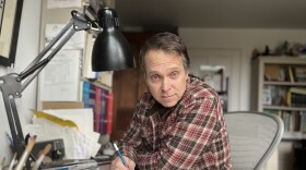 A man with short salt and pepper hair is wearing a plaid button-down shirt and drawing on a pad while sitting at a desk in an office.