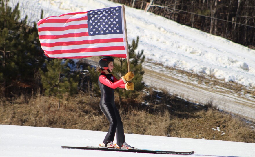 A girl carries an American Flag as she skis during the Itasca Ski Club's tournament opening ceremonies at Mount Itasca on Feb. 18, 2024.