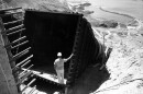 A man in a hard hat stands inside a massive dry pipe, looking down to a reservoir downhill 