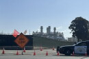 A road closed sign, police car, and traffic cones are shown in front of industrial smoke stacks. 