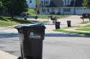 A black city of Columbia waste roll cart stands at the end of a driveway in a residential neighborhood. More roll carts and houses are visible in the background.